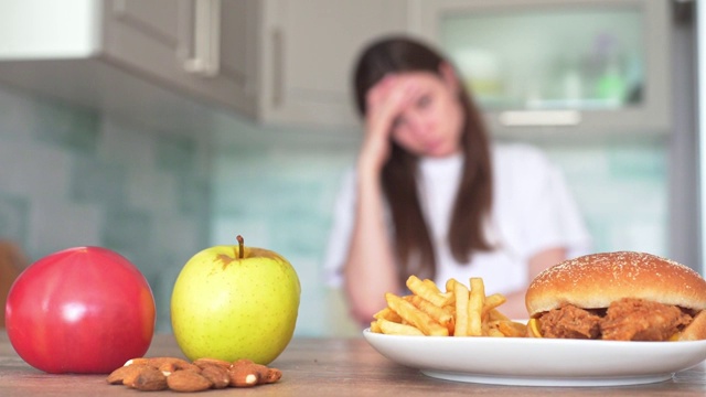 不健康食品和健康食品之间的选择。汉堡和薯条前面是苹果，西红柿和坚果。苗条的女孩在厨房里试图做出艰难的食物选择视频素材