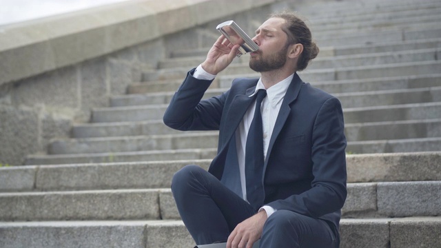 一个抑郁的男人坐在城市的楼梯上喝着酒瓶里的酒。沮丧的白人雇员失去了他的工作。患有抑郁症的年轻悲伤的商人。视频素材