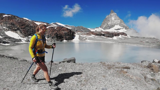 慢镜头——女登山运动员拿着登山杆在湖边行走，马特洪峰是背景视频素材