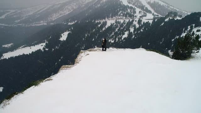 从山峦环绕的悬崖边回来的情侣在雪下拥抱。美丽的雪景与山脉遍布地平线- 4K视频素材