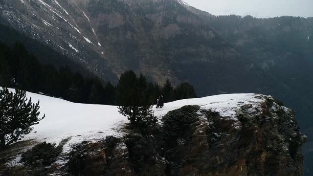 夫妇俩下着雪，走在悬崖边上，四周群山环绕。美丽的雪景与山脉遍布地平线-鸟瞰图与无人机4K视频素材