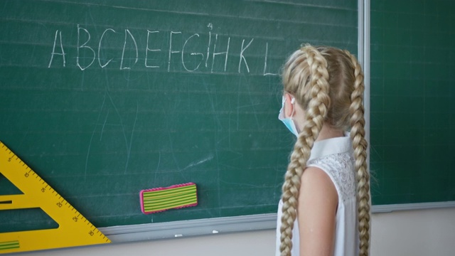 戴着医用口罩的女学生在教室里用粉笔写英语字母，小学生在学校的黑板旁看着摄像机视频素材