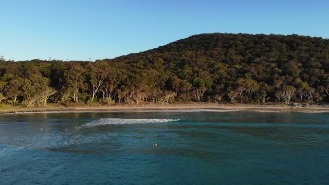 一个阳光明媚的夏日夜晚，努萨岬的茶树湾。海浪在蓝色的海水中劈开，冲浪者划桨而出。阳光在国家公园投下阴影视频素材