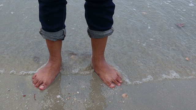 一个女人站在沙滩上，被海浪拍打着。海浪和风吹在沙滩上。在晚上的时间。视频素材