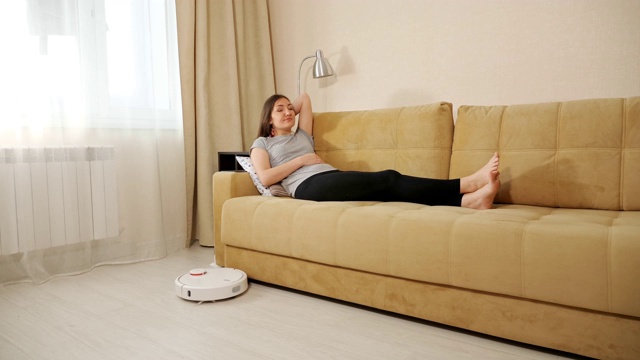 快乐的女人躺在沙发上，机器人清洁工打扫地板视频素材