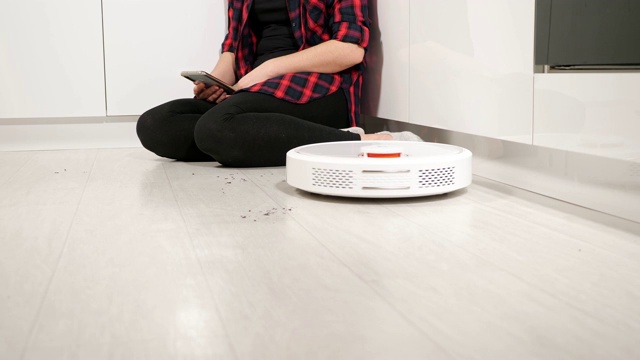 一个拿着手机的陌生女人坐在地板上，而机器人在清扫地板视频素材