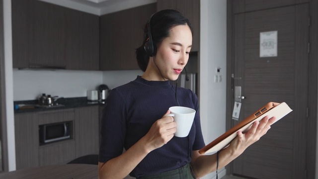 亚洲女性戴着耳机和使用数字平板电脑视频会议和会议，在家里的厨房喝咖啡视频素材