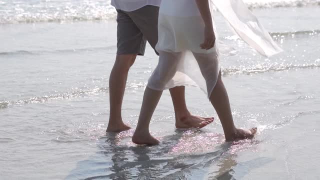 微笑的年轻亚洲夫妇男人和女人手牵着手一起在夏天的海滩上散步的慢动作。幸福的家庭，丈夫和妻子放松，享受浪漫的暑假。视频素材