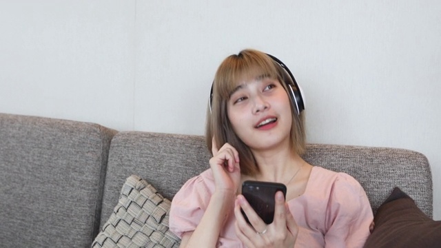 亚洲女性喜欢在家里放松的时候戴着耳机在手机上听音乐视频素材