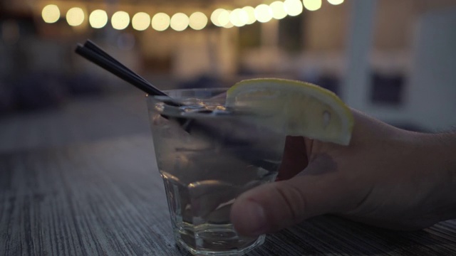 白色的皮革男性的手带来一个低杯，雾蒙蒙的透明冷饮一杯一片柠檬，两根饮用吸管和冰块到餐厅的木桌。饮料和鸡尾酒主题。视频素材