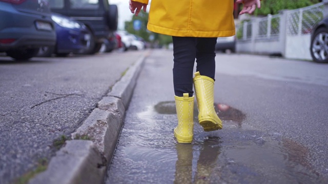 穿着黄色胶靴和雨衣的年轻女孩走过街道上的水坑视频素材