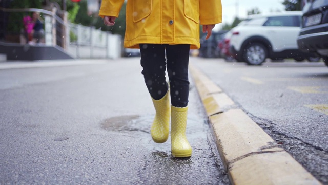 穿着黄色胶靴和雨衣的年轻女孩走过街道上的水坑视频素材