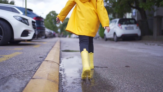 可爱的孩子穿着黄色胶靴和雨衣在街上的水坑里跑视频素材