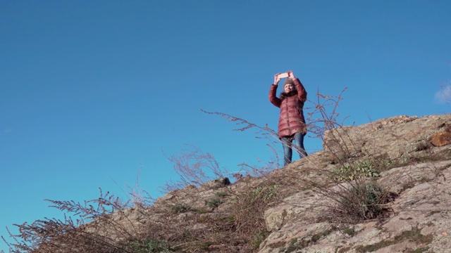 一位年轻女子正在徒步旅行。她爬上山顶，用智能手机拍下了美丽的风景。视频素材