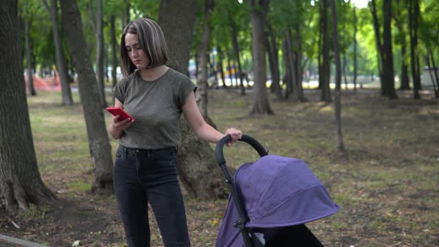 一名妇女在户外摇着婴儿车里的婴儿，一边看手机视频素材