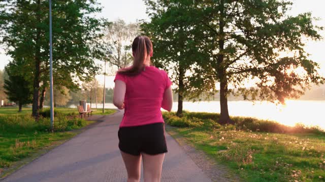 女性慢跑者在马路和街道上跑步，公园里有树。在夏天的早晨慢跑的女人。跑步者在阳光明媚的大自然中进行户外有氧运动。视频素材