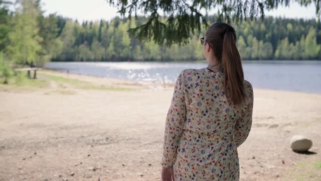 在海滩上散步的女人。穿着花裙子和夏装的女士。在芬兰享受周末或假期的漂亮女孩。湖泊景观与水和森林。在大自然中放松。视频下载