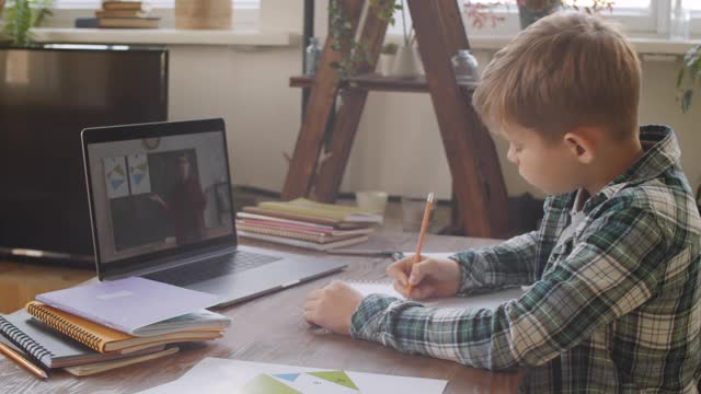 男孩在书桌上写作，同时在家里通过笔记本电脑视频电话学习视频素材