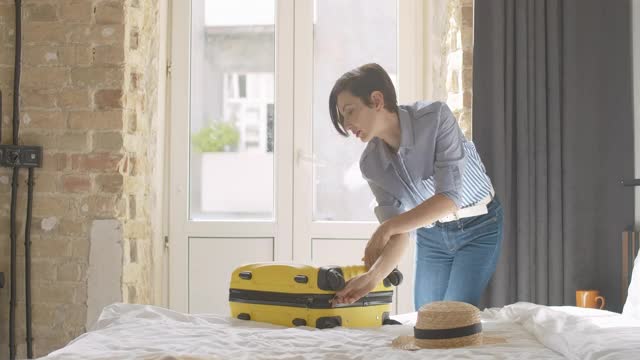 自信美丽的女人拉链黄色旅行袋和离开酒店房间或卧室行李。年轻迷人的白人女游客带着行李在室内。视频素材