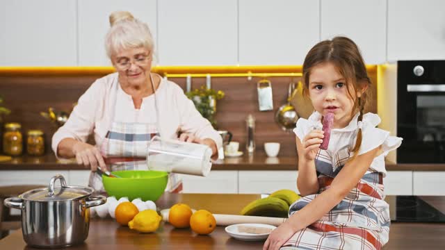 奶奶把面粉和鸡蛋放在碗里，用搅拌器搅拌，小孙女坐在桌子上吃糖果，在现代厨房里做饼干视频素材