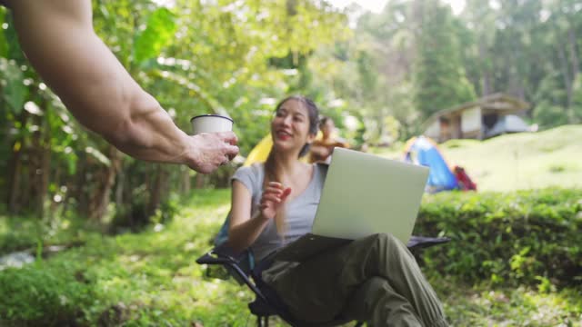 美丽的亚洲女人使用笔记本电脑与互联网在森林营地户外工作的慢镜头。一对幸福的夫妇坐在小溪对面的椅子上，一起喝着咖啡。具有科技理念的户外生活方式。视频素材