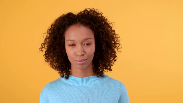 可爱谦逊的非裔美国女人看着照相机在蓝色毛衣。人在孤立的黄色背景。4 k视频素材