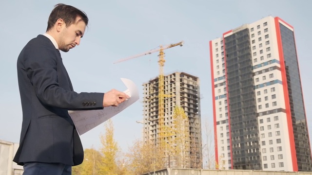 一位年轻的男建筑建筑师正在检查大楼的施工情况。一个工程师看着图纸，背景是街道上的建筑，背景是一座未完工的高层建筑视频素材