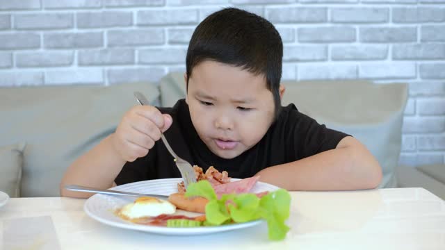 一个亚洲小男孩在家里吃着煎蛋、培根、土豆和香肠的早餐。视频素材