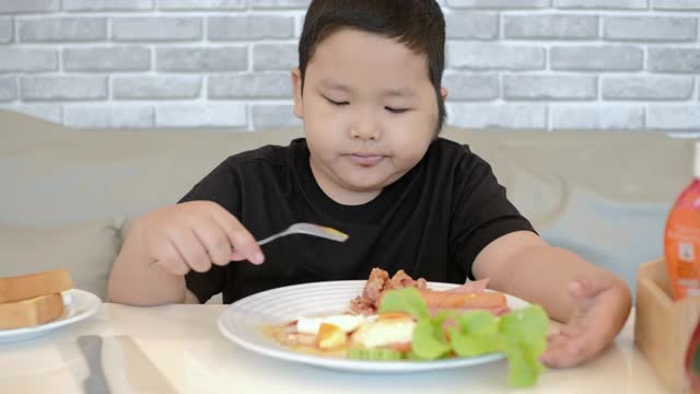 一个亚洲小男孩在家里吃着煎蛋、培根、土豆和香肠的早餐。视频素材