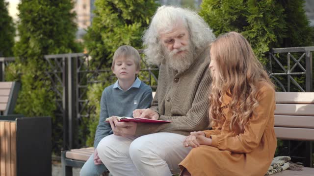 角度视图的智能男性退休人员解释书的孙子在阳光灿烂的一天户外。自信快乐的白人祖父的肖像为男孩和女孩坐在长凳上阅读。视频素材