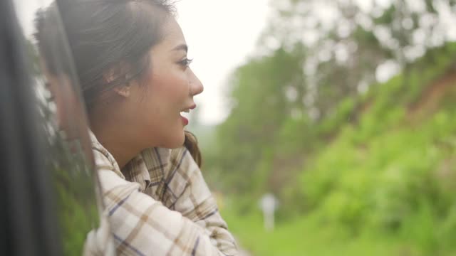 慢动作的快乐年轻美丽的亚洲女人旅行者坐在汽车与拉她的脸和手在雨天的车窗。漂亮女孩喜欢和享受户外生活方式和假期的乐趣。视频素材