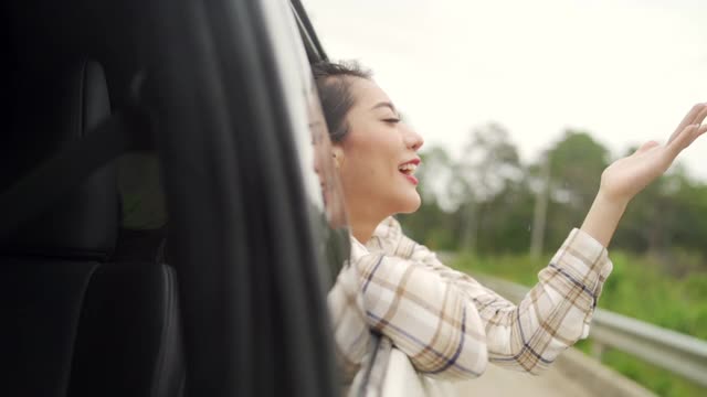 慢动作的快乐年轻美丽的亚洲女人旅行者坐在汽车与拉她的脸和手在雨天的车窗。漂亮女孩喜欢和享受户外生活方式和假期的乐趣。视频素材