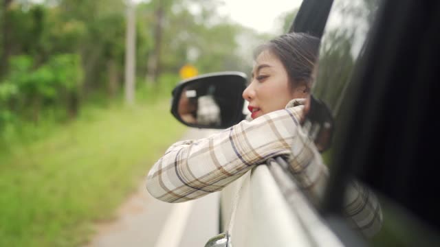 慢动作的快乐年轻美丽的亚洲女人旅行者坐在汽车与拉她的脸和手在雨天的车窗。漂亮女孩喜欢和享受户外生活方式和假期的乐趣。视频素材