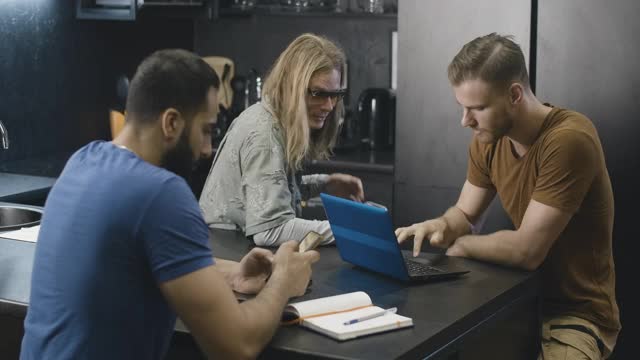 三个积极的大学生坐在宿舍厨房里聊天。欢快的年轻人用智能手机和笔记本电脑上网聊天。电影4k ProRes总部。视频素材