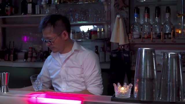 4K专业调酒师在水晶鸡尾酒杯中加入冰块，用于夜总会酒吧调制鸡尾酒。男调酒师调酒师制作酒精饮料与烈酒架子的背景。视频下载