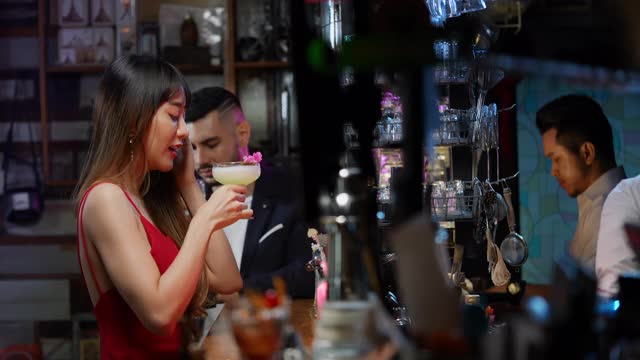 4K微笑美丽的亚洲女人站在吧台，享受着酒吧调酒师提供的美味鸡尾酒。男调酒师用装饰性的鸡尾酒杯准备混合酒精饮料。视频下载