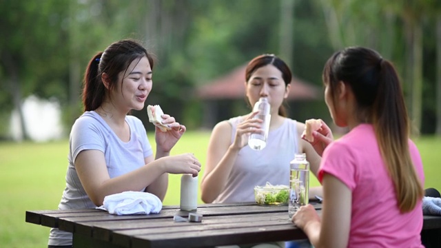 3个亚洲中国年轻女子在公园休息后早上锻炼吃健康的食物和饮料在公园长椅视频素材