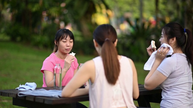 3个亚洲中国年轻女子在公园休息后早上锻炼吃健康的食物和饮料在公园长椅视频素材