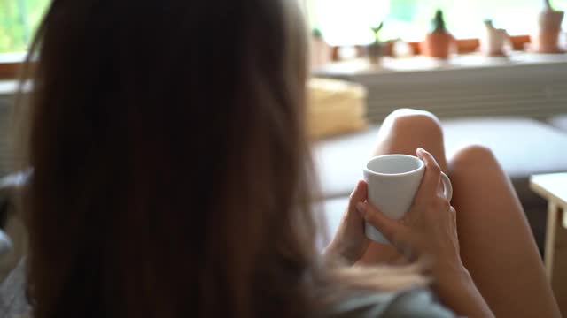 滑右图，轻松的年轻女子拿着咖啡杯坐在沙发上。高质量4k镜头视频素材