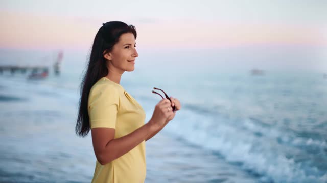 微笑的女人穿着黄色的衣服戴着太阳镜欣赏海景。用RED摄像机拍摄4K视频素材