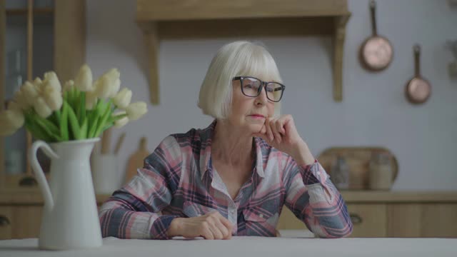 有趣的70年代女人戴着眼镜，用手指表示胜利的手势，坐在白色郁金香的餐桌上，看着镜头。快乐的资深金发女人在镜头前做V手势和舌头，微笑着。视频素材