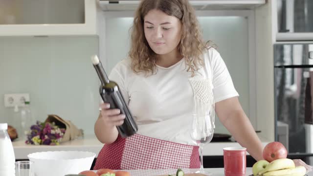 深色头发的大个子白人女人打开瓶子，把红酒倒进玻璃杯。中景肖像微笑的大码女士品尝昂贵的饮料在家里的周末。酒精的概念。视频素材