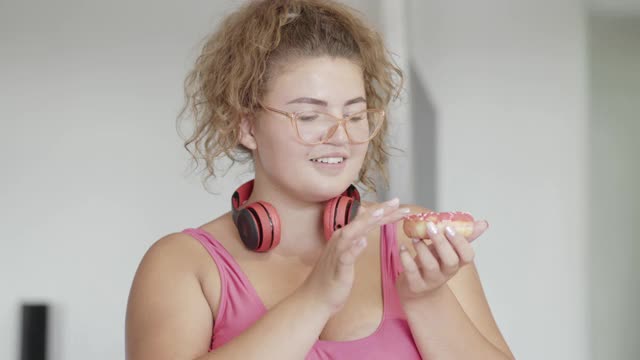 大码卖弄风情的女人品尝美味的甜甜圈和微笑的镜头。特写的快乐的白人大身材女士享受甜美可口的面包房室内。调情和生活方式。视频素材