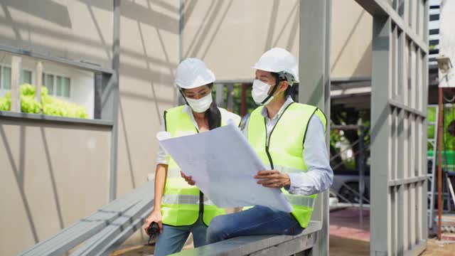 亚洲工人在建筑现场佩戴防护口罩。男女工程师拿着图纸在施工现场工作，讨论项目。视频素材