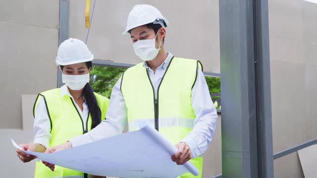 亚洲工人在建筑现场佩戴防护口罩。男和女工程师拿着图纸在施工方讨论项目。视频素材