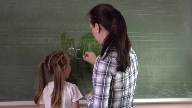 一个年轻的老师和一个小女孩正在黑板上写“学校”这个词。视频素材