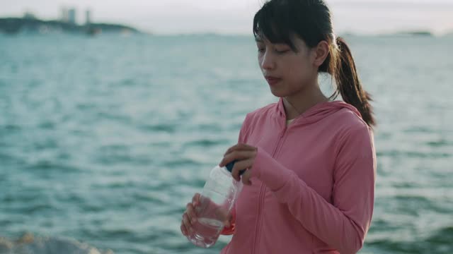 女跑步者站在海边喝水。视频素材