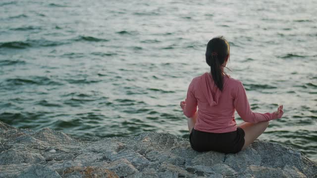 年轻美丽的运动女孩训练冥想坐在石头海边，而日落。视频素材