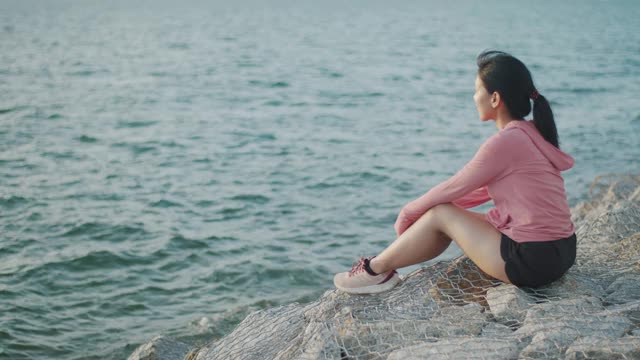 年轻女子跑步者坐在海边的石头上，从锻炼中休息，欣赏海滩日落的景色。视频素材