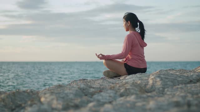 亚洲运动女性沉思坐在海边的石头日落。视频素材